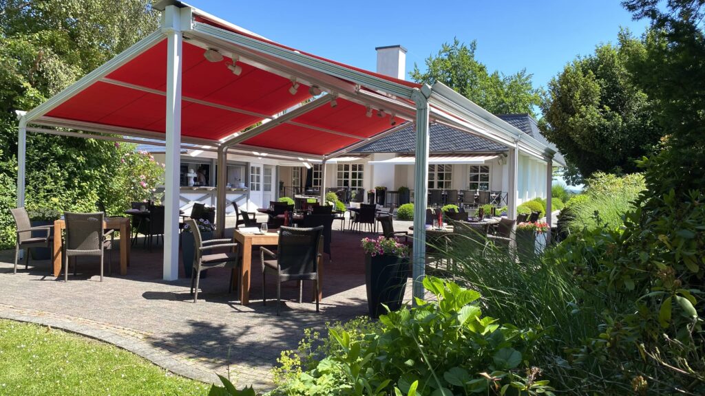 Terrasse eines Restaurants mit roten Markisen und Tischen im Freien, umgeben von Pflanzen und Bäumen.