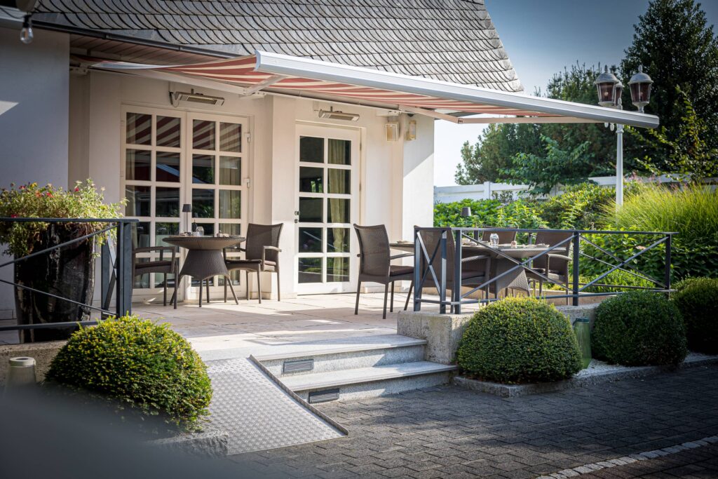 Terrasse mit Tisch und Stühlen vor einem Gebäude mit großen Fenstern und einem schattenspendenden Sonnenschutz.