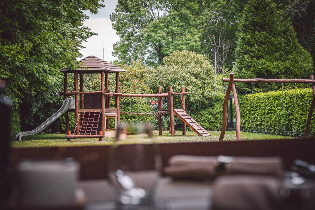 Spielplatz mit Holzspielgerüst, Rutsche und Schaukeln in einem Gartenbereich.