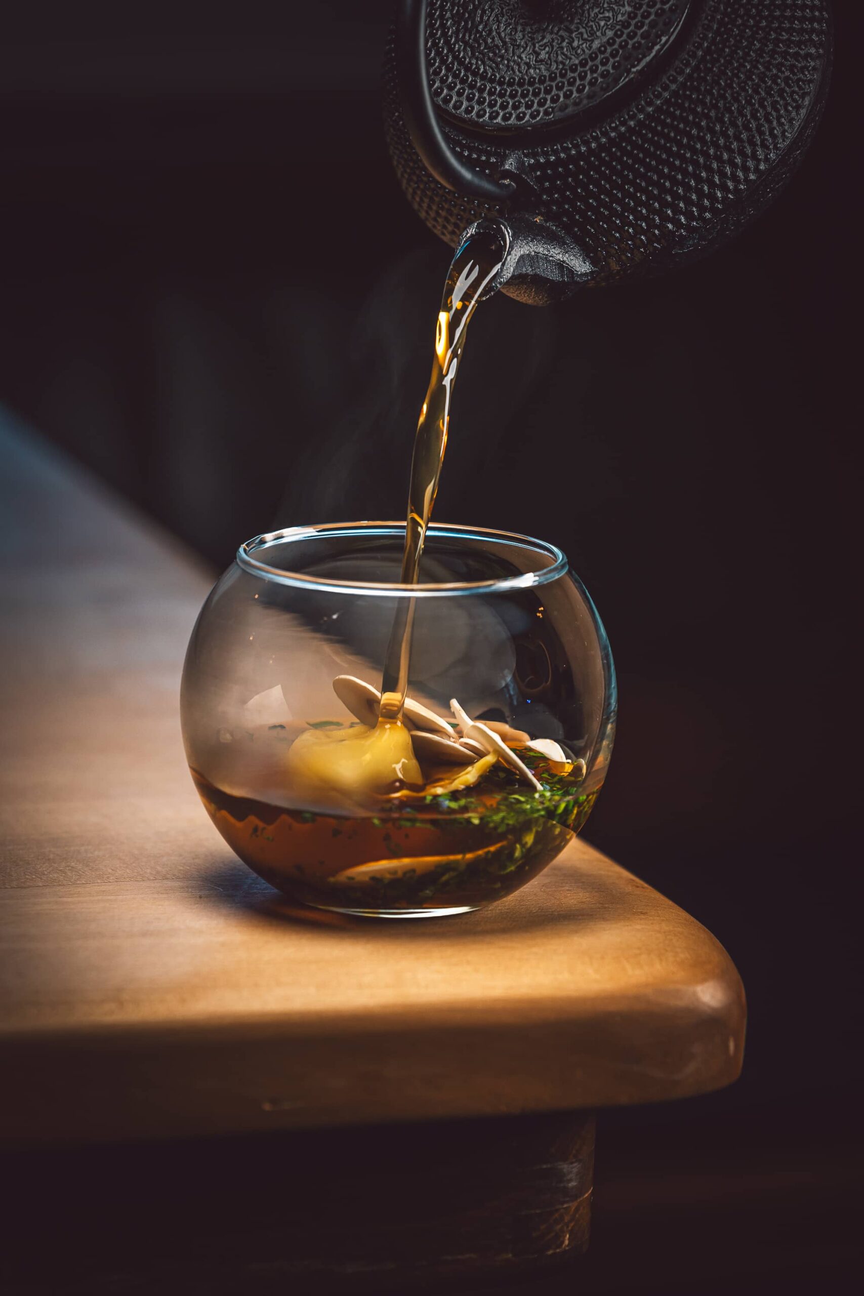 Eine Teekanne, die Tee in ein Glas mit Kräutern und einer Holzlöffel gießt.