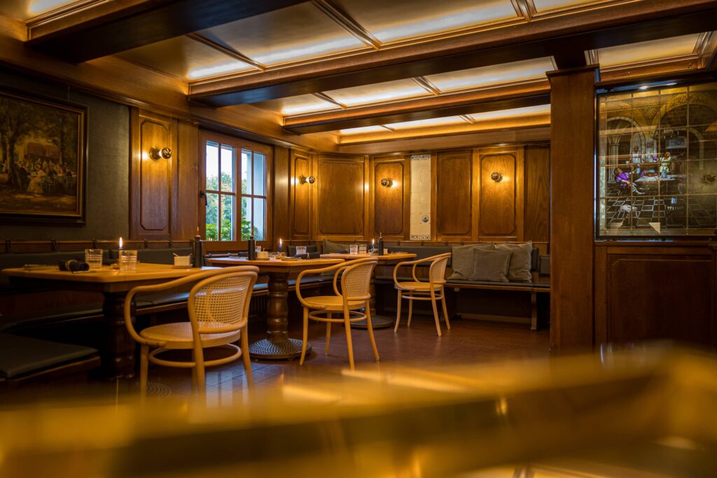 Innenansicht eines Restaurants mit Holzmöbeln, Tischen und Stühlen, beleuchtet von Wandleuchten.